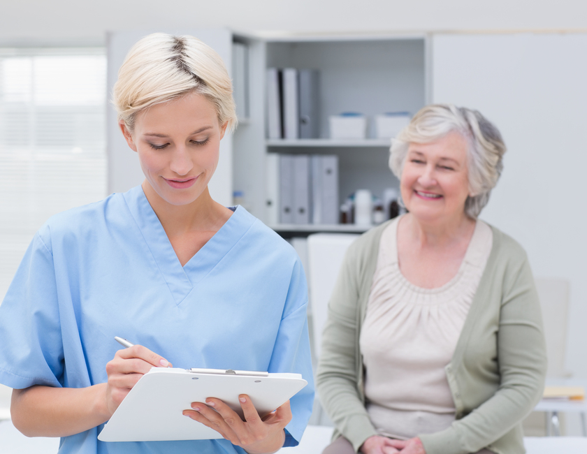 Female nurse writing on clipboard while senior patient sitting in clinic