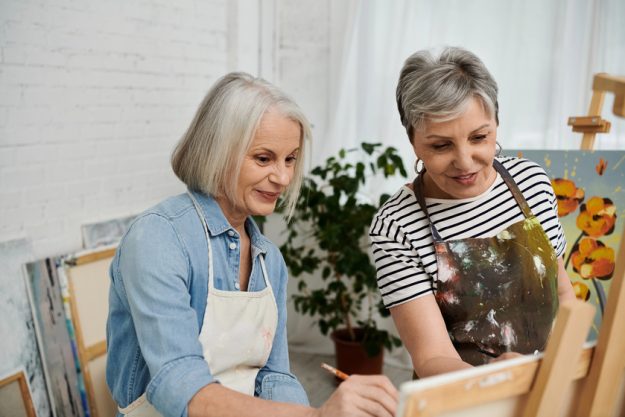 Two senior women in an art studio, one painting on a canvas while the other looks on, sharing a moment of creative connection.