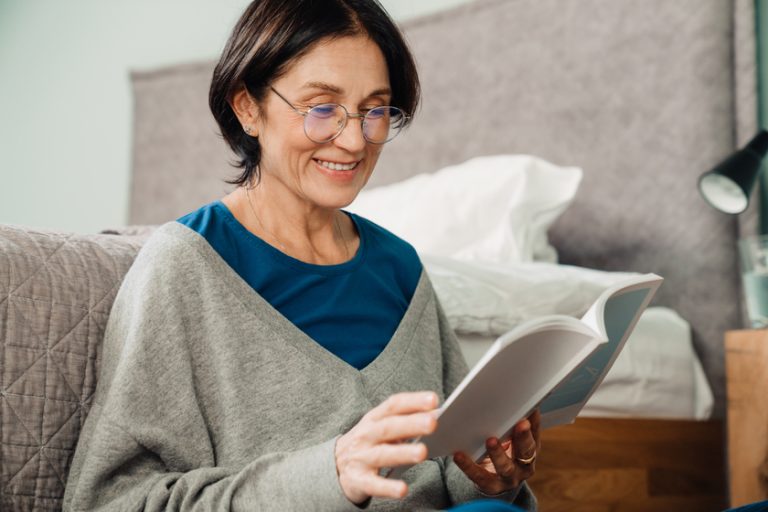 White mature woman in eyeglasses reading book while sitting on floor