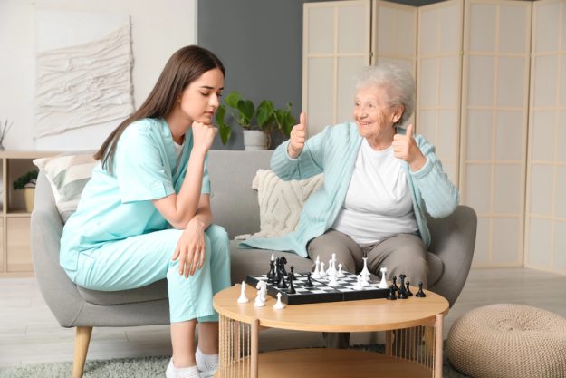 Young caregiver with senior woman playing chess at home