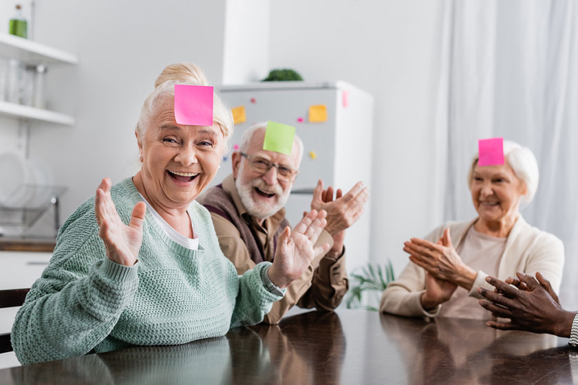 Excited multicultural senior friends sticky notes foreheads playing guess who Excited multicultural senior friends sticky notes foreheads playing guess who