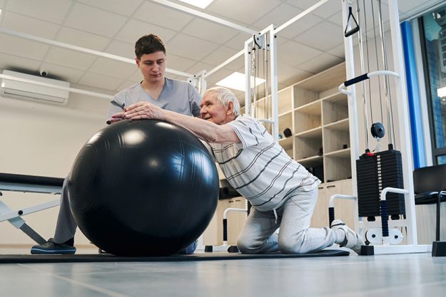 Pensioner standing on knees while putting arms on exercise ball